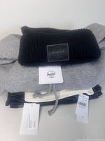 Four clothing items neatly stacked showing tags and brand labels. Herschel black knit toque on top, gray TNA hoodie beneath, H&M tank top visible, and American Eagle jeans at the bottom.