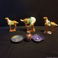 Front view of three porcelain camel figurines behind two cloisonné plates and one vase on black cloth background.