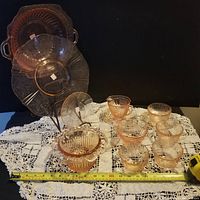 Wide shot showing multiple pink depression glass items including cups, bowls, and plates spread on a crocheted tablecloth with a measuring tape across the bottom for scale.
