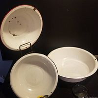 Three vintage enamel ware bowls arranged on black surface with one hanging vertically on a display stand, showing wear, chipping and rust spots.