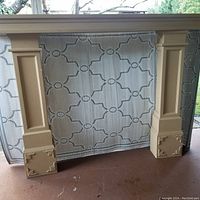 Three-piece beige painted wooden fireplace mantle set with decorative floral corner accents on bases, shown leaning against fabric backdrop outdoors on concrete surface.