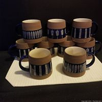 Full set of 11 pottery cups grouped on a white cloth, showing the painted designs and blue handles.