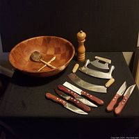 Photo showing all items together: large segmented wooden bowl, wooden ladle and spatula, wooden pepper mill, mezzaluna knife, curved blade knife, six wooden handled steak knives.