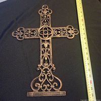 Front view of large metal crucifix showing intricate scrollwork and floral center on black background with measuring tape for scale.