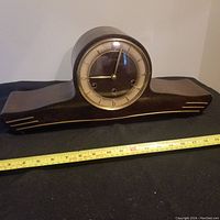 Front view of a vintage wooden mantle clock showing curved dark wood body with cream dial and gold numerals, positioned beside a measuring tape indicating approximately 17 inches in width.