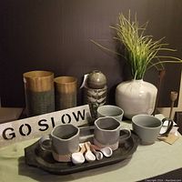 Wide view showing the majority of the lot arranged on table with cups, trays, sign, ceramic vase with faux grass, metal vases and spoons.