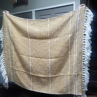Photo of a beige throw blanket held up by two hands showing white vertical stripes and white fringe on edges.