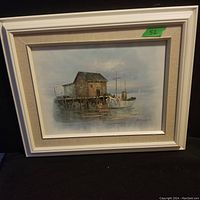 Framed oil painting showing fishing boat docked near wooden building on reflective water.