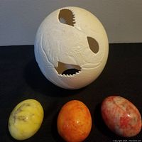 Large carved ostrich egg with rhinoceros design and three smaller polished marble eggs in yellow, orange, and red tones on black surface.