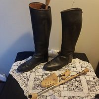 Side view of the pair of dark brown vintage riding boots shown standing on lace tablecloth with two wooden boot stretchers in front.
