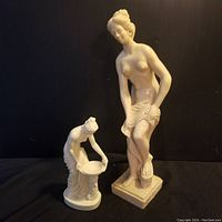 Two nude female figurine sculptures on a black background, showing size difference and poses of each statue.