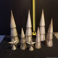 Seven cone-shaped silver metal tree decorations and two star-shaped toppers arranged on black background with measuring tape.