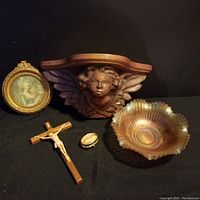 Five vintage items arranged on dark background including a carved wooden angel wall shelf, framed miniature portrait, wooden crucifix, small locket, and decorative brass bowl.