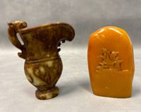 Two miniature carvings: a brown Shoushan stone pitcher and an orange rectangular stone with carved figures.