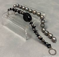 Both bracelets displayed on clear riser showing pearl and black beads with silver clasps.