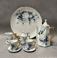Photo showing entire dollhouse tea set arranged with plate, teapot, sugar bowl, creamer, two teacups and two saucers. Floral white rose and purple flower design with gold trim on all items.