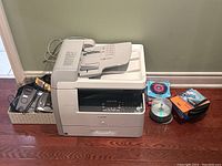 Front and side view of Canon multifunction printer with accessories arranged beside it on floor under wall with light greenish paint.