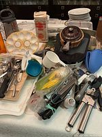 Various kitchen and baking tools including white egg holder, metal grater, brown ceramic pot with lid, black and blue oven mitts, measuring cups, and various metal utensils