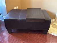 Photo of the dark brown leather ottoman with a black wooden removable table surface on top, showing the full ottoman and table surface from the side.