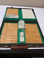 Open brown carrying case revealing four compartments filled with many Mahjong tiles and a central section with dice and scoring sticks in plastic bags.