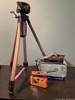 Full view of Optex tripod with Polaroid Joycam camera on top and original box, along with two disposable cameras in front.