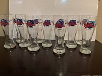 Set of 11 pilsner beer glasses with Planet Hollywood logos from Canadian and US cities lined up on a dark surface, front view.