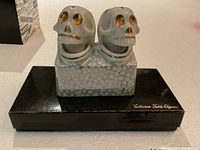Front view showing two skull-shaped shakers on patterned ceramic base, placed on black box.