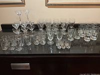 Full set of vintage etched glasses on black countertop showing multiple types including stemmed glasses, shot glasses, and tumblers arranged in rows.