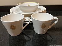 Photo showing two white coffee mugs with gold Royal Naval Air Service emblem on front, with rimmed bowl and lunch plates stacked behind.