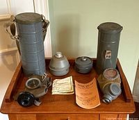 Two Belgian WW2 gas masks displayed on a wooden surface with their original metal storage cans and instruction manuals. The masks show signs of rubber cracking and aging.