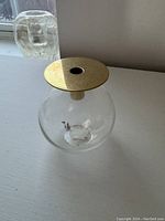 Front angled view showing the spherical clear glass vase with gold removable top and hole for a bud, placed on a white surface with a textured glass object in the background.