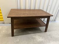 Front facing view of rectangular wooden coffee table with lower shelf, brown wood grain finish, standing on concrete floor near metal wall.