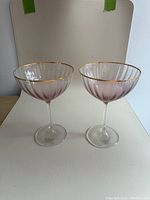 Two pink tinted ribbed cocktail glasses with gold rims and clear stems sitting on a white surface, photographed from front angle.