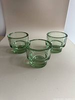 Three green glass tealight candle holders placed on a flat white surface with a white backdrop. Each holder is cylindrical with thick glass walls and a solid base.