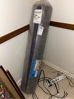 Full view of the rolled gray textured rug underlay wrapped in plastic, showing packaging labels and a blue product label with installation room example.