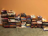 Photo showing long rows and stacks of assorted books set on carpet against a plain wall, includes paperbacks and hardcovers.