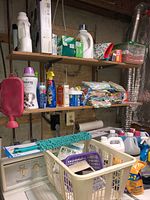 Photo of multiple shelves in basement holding assorted cleaning supplies including bottles of detergent, spray bottles, folded cloths, and plastic baskets.