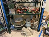 Full view of a green metal shelf holding a faux lavender plant in a ceramic pot, fruit-patterned tray, stacked bowls and plates, two gray squirrel garden statues on bottom shelf.