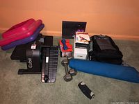 Wide view of lot including Cubii elliptical, dumbbells, dumbbell box, ankle weights, DVDs, yoga mat, inflatable rings, and backpack on carpeted floor.