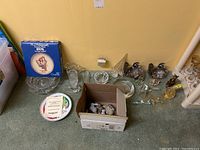 Photo showing all items including the boxed Hummel plate, cut glass bowls, various glass bird and fish figurines, and ceramic figurines in a cardboard box, arranged on carpeted floor.