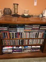 Photo showing multiple shelves with CDs, DVDs, and cassettes packed tightly in wooden shelving unit, few decorative ceramic items on top surface