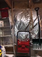 Photo showing Werner 6 foot aluminum step ladder, red folding stepstool, and green snow shovel with wooden handle and black D-grip handle, all located in basement against silver insulating material on wall.