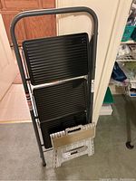 Photo showing the folding three step stool with black ribbed metal steps and the gray plastic one step stool placed in front.