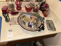 Overview of collection including red glass pieces, perfume atomizers, candle holder with candle, woven tray filled with nail accessories, ceramic owl dish, and watercolor palette on a white countertop.
