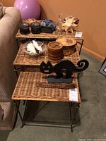 Three wicker nesting tables stacked inside each other, arranged vertically. Top table smallest, bottom largest. Tables natural wicker color with black metal legs and frame. Photo taken to show condition and nesting nature.