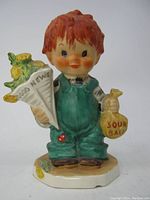 Front view of figurine showing child holding "Good News" newspaper and "Sour Balls" bag.