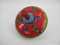 Top-down view of the vintage Buck Rogers spinning top toy showing the red handle and lithographed design with astronauts and rockets on a red background.