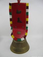 Front view of vintage brass cowbell with detailed embossing, Swiss crest, and red leather strap with colorful fringes and metal emblems.