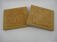 Two wooden plaques with round-edged rectangular shapes showing raised carvings of stylized Indigenous designs on flat wooden background.
