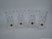 Front view of four clear Karl Ittig handmade shot glasses showing blue dots on upper portion and amber dots near base.